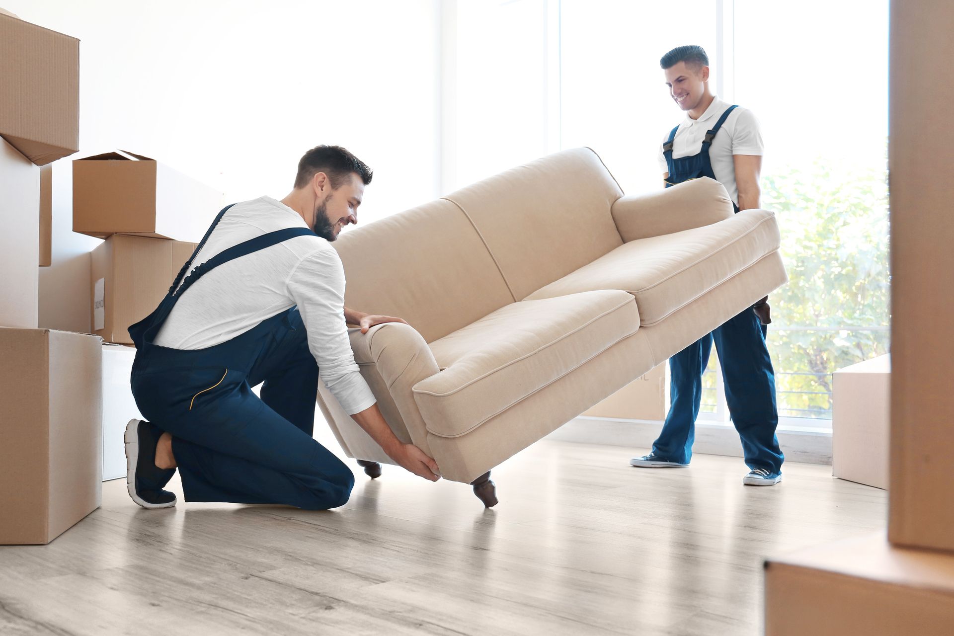 Zwei Männer in blauer Arbeitskleidung heben ein hellbeiges Sofa in einem Raum mit Umzugskartons und hellem Holzboden.